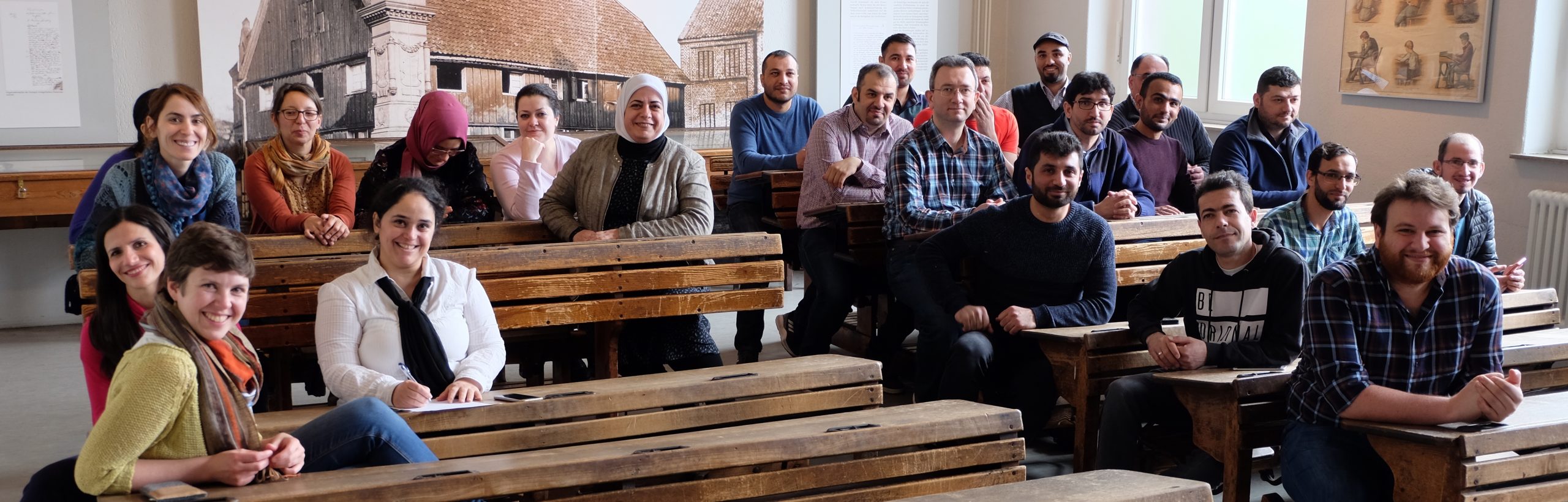 Eine Gruppe erwachsener Menschen sitzen auf historischen Schulbänken.